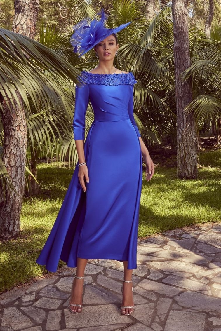 The image showcases a woman positioned centrally on a stone pathway in an outdoor garden. She is wearing a fitted, long-sleeve blue gown with intricate embellishments around the neckline, flowing into a slightly flared skirt. She stands confidently, holding one hand at her side and the other slightly raised. Her wide-brimmed blue hat, decorated with feather-like details, complements the dress. The background includes several tall trees and dense palm foliage, enhancing the bright, natural ambiance under a c